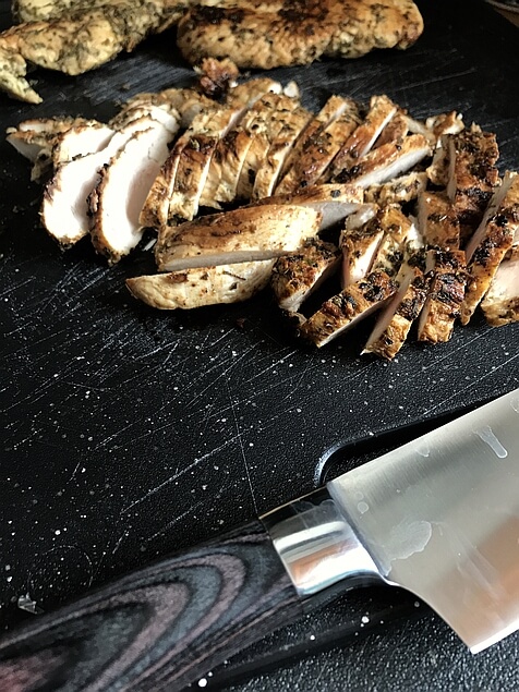 Cutting board and a chef's knife help frame this picture of freshly sliced, perfectly cooked, juicy chicken breast. Two other breasts sit on the back of the cutting board behind the sliced ones, slightly out of focus.