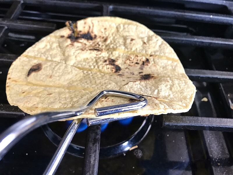 Tortilla held with tongs, being warmed and toasted directly over the burner of a gas stove.