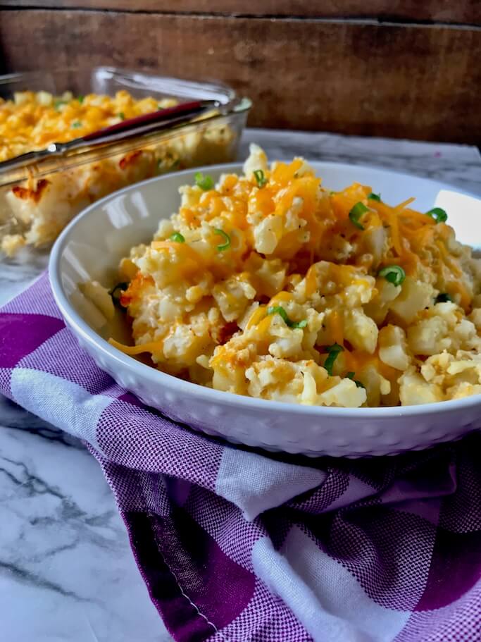 Side view of a bowl of freshly-cooked Skinny Cheesy Potatoes. The entire casserole dish is visible in the background but with a very noticeable scoop missing, giving us a look at the warm layers of gooey goodness inside!