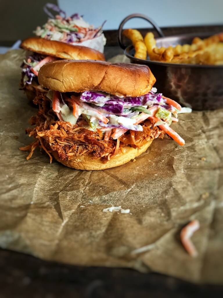 Crockpot Pulled BBQ Chicken sandwich on parchment paper with a bowl of coleslaw and fries in the background
