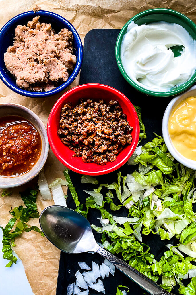 Ingredients: shredded lettuce, spoon, bowls of seasoned ground beef, salsa, refried beans, greek yogurt, cheddar cheese