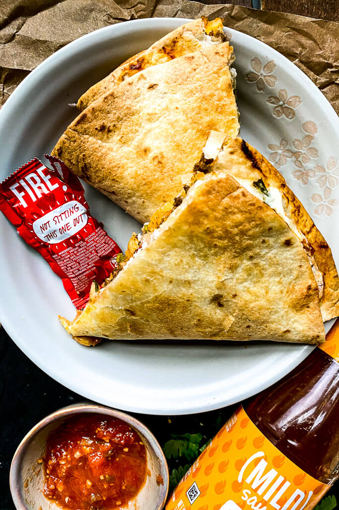 taco crunchwrap on a plate with a small bowl of salsa, taco bell red hot sauce packet, and a bottle of mild taco sauce