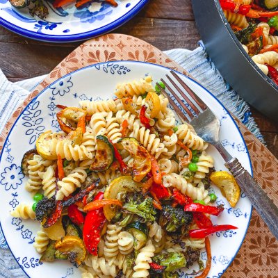pasta primavera with roasted vegetables on a blue and white plate with fork and cutting board in background