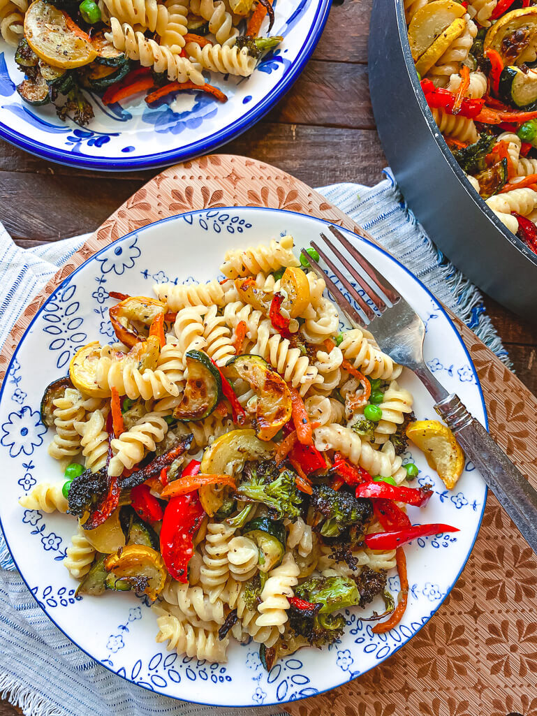 pasta primavera with roasted vegetables on a blue and white plate with fork and cutting board in background