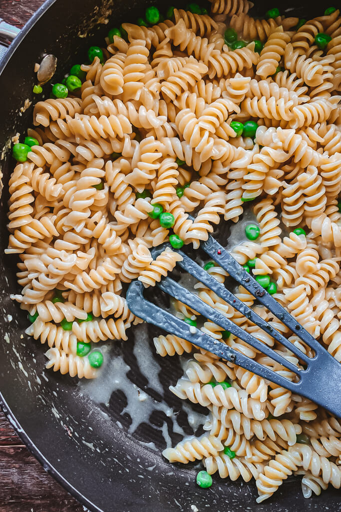pan with cooked rotini a bright green peas being stirred with a blue spatula 
