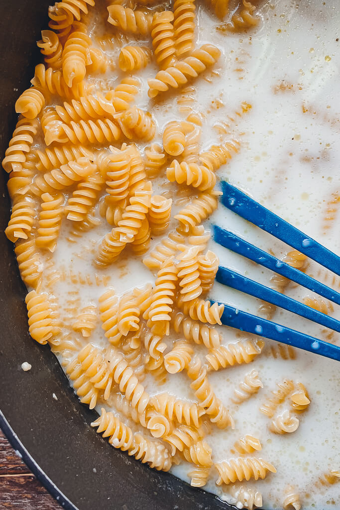 pan with rotini noodles and milk being stirred with a blue spatula 