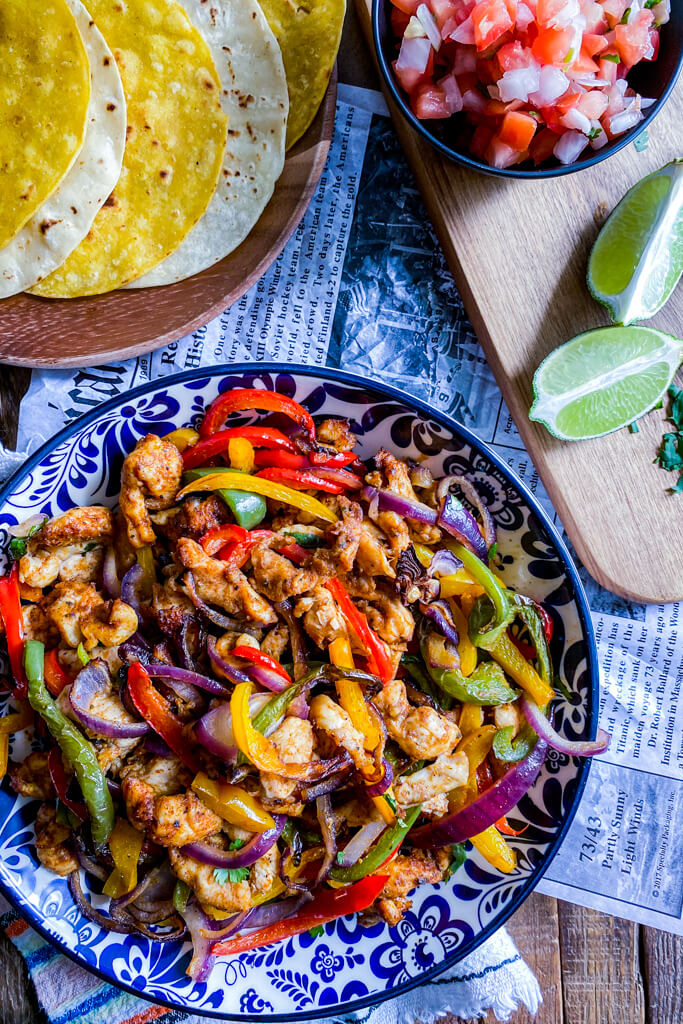 chicken fajitas in blue and white bowl yellow and white corn tortillas on wooden plate tomatoes and onion in black bowl and lime on a wooden cutting board