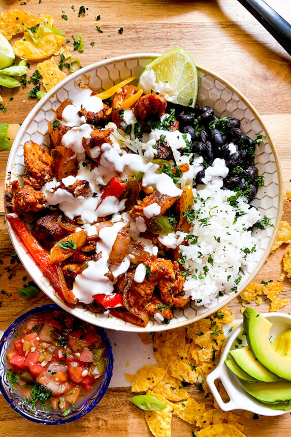 pico de Gallo and sliced avocado in small bowl, large white bowl filled with grilled chicken and peppers, white rice, black beans, lime wedge, sour cream sauce, and sprinkled with cilantro
