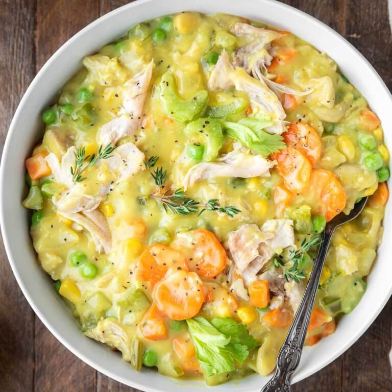Creamy chicken pot pie soup in a white bowl with shredded chicken, carrots, peas, corn, and celery, garnished with fresh thyme.