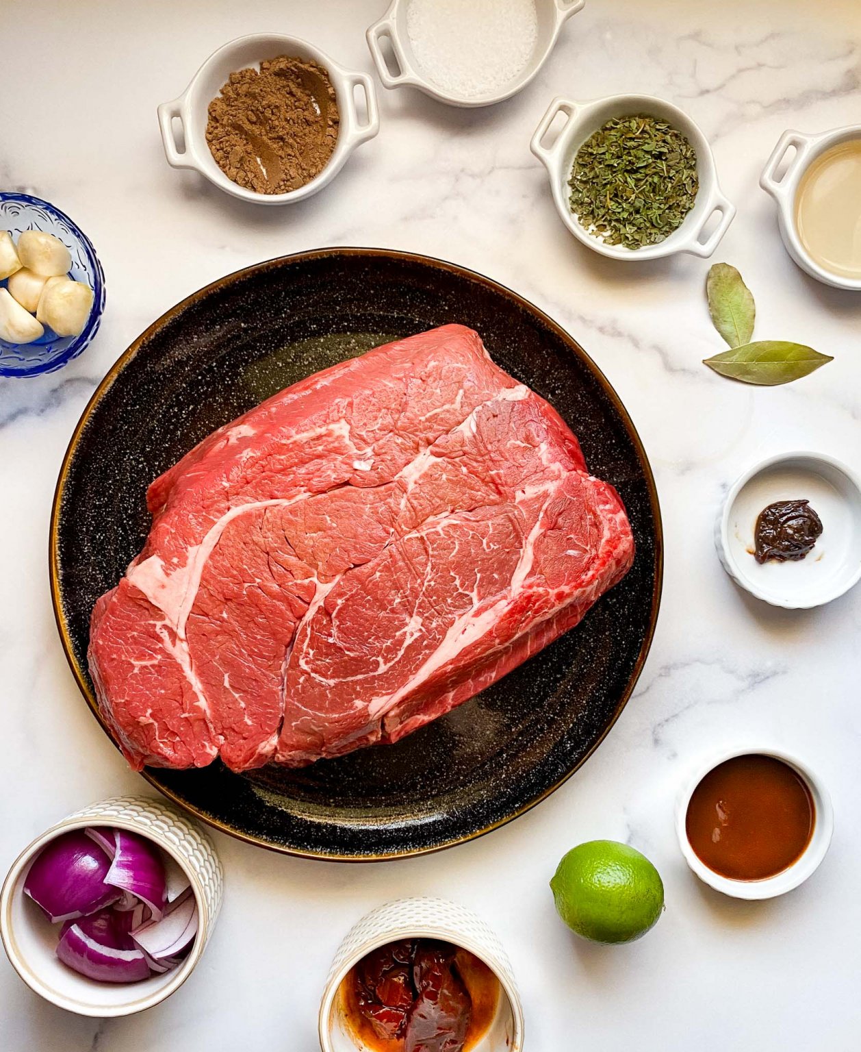 photo of ingredients for recipe large plate with a raw chuck roast surrounded by small bowls of garlic cloves, ground cumin, kosher salt, dried oregano, bay leaves, apple cider vinegar, better than bouillon, adobo sauce, lime, chipotle peppers, red peppers