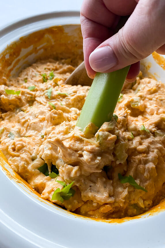 buffalo chicken dip in slow cooker with hand scooping up some with a piece of celery