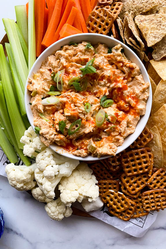 big platter with fresh carrots, celery, and cauliflower next to pretzels and tortilla chips with a big bowl of buffalo chicken dip centered