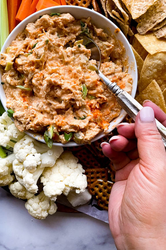 platter of celery, carrots, tortilla chips, cauliflower, and pretzels with a big bowl of buffalo chicken dip in the center with a spoon inside of it and a hand grabbing the spoon about to scoop it out of the bowl.