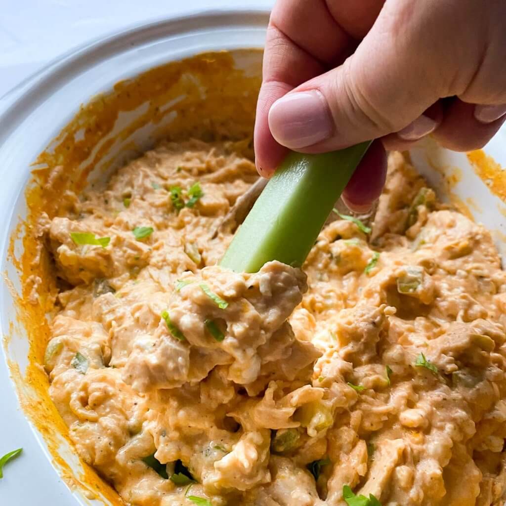 white crockpot full of cooked buffalo chicken dip with a hand holding a piece of celery and dipping it into the dip