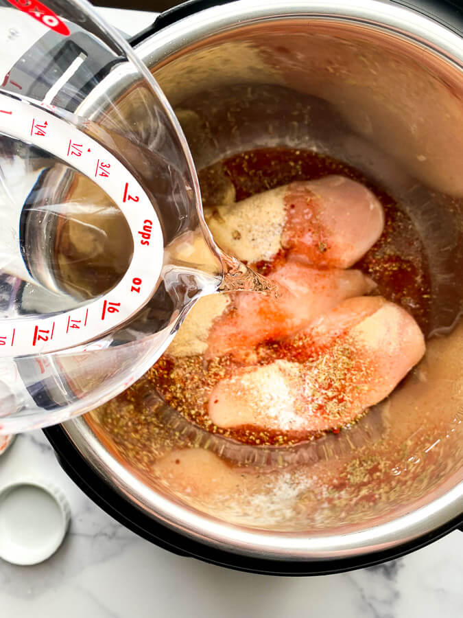 boneless skinless chicken inside instant pot with spices and water being poured onto the chicken from a measuring cup
