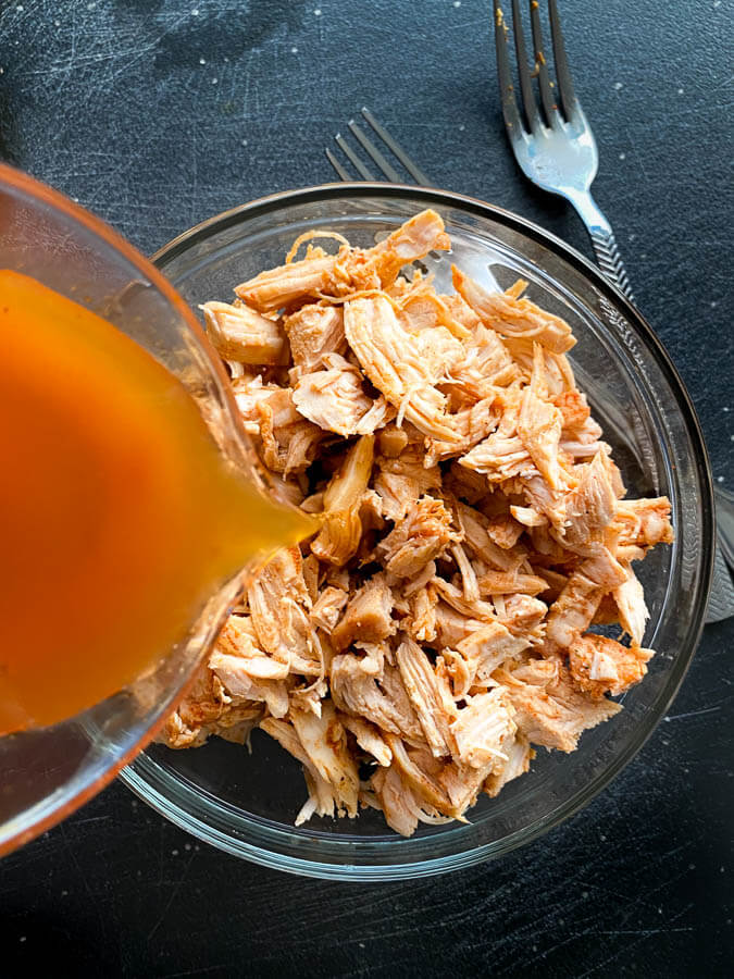 Cooked and shredded chicken in a glass bowl with chicken broth being poured into the bowl and forks on the side