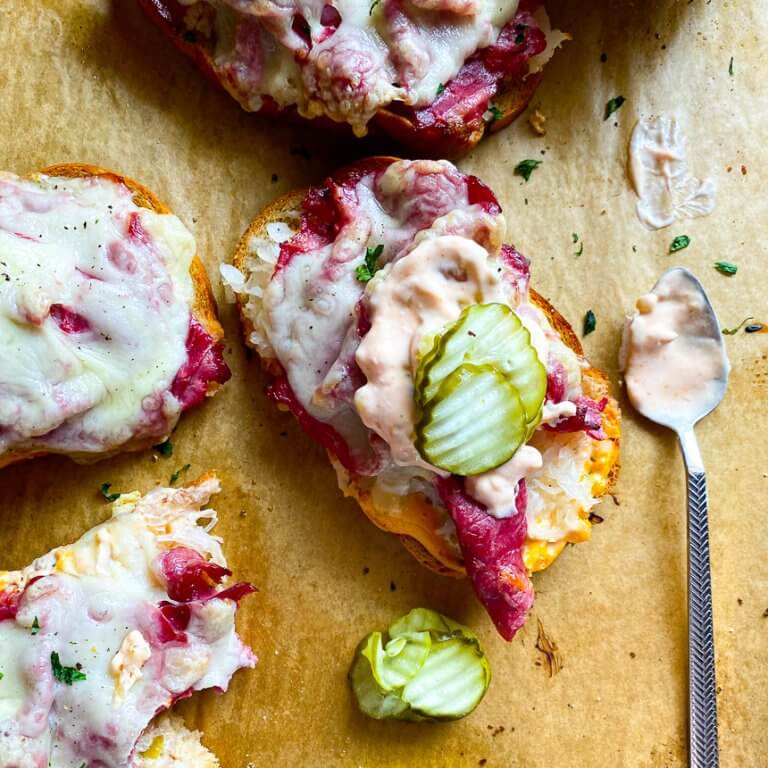 Open Faced Reuben Sandwich with a spoon on the side with thousand island dressing and pickles next to it