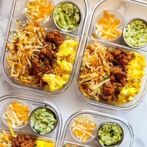 clear meal prep containers filled with scrambled eggs, turkey taco meat, shredded hash browns and small bowls of guacamole and shredded cheese