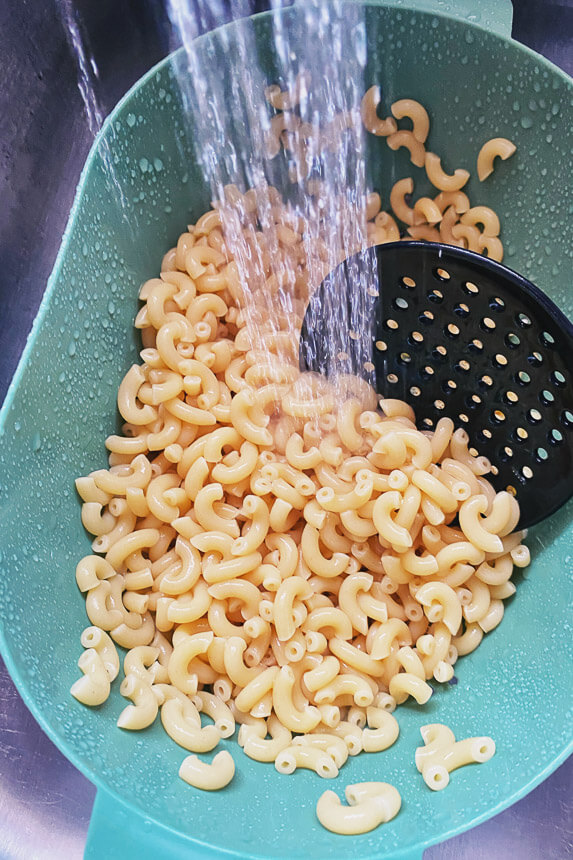macaroni noodles in a green colander being sprayed with cold water in a sink