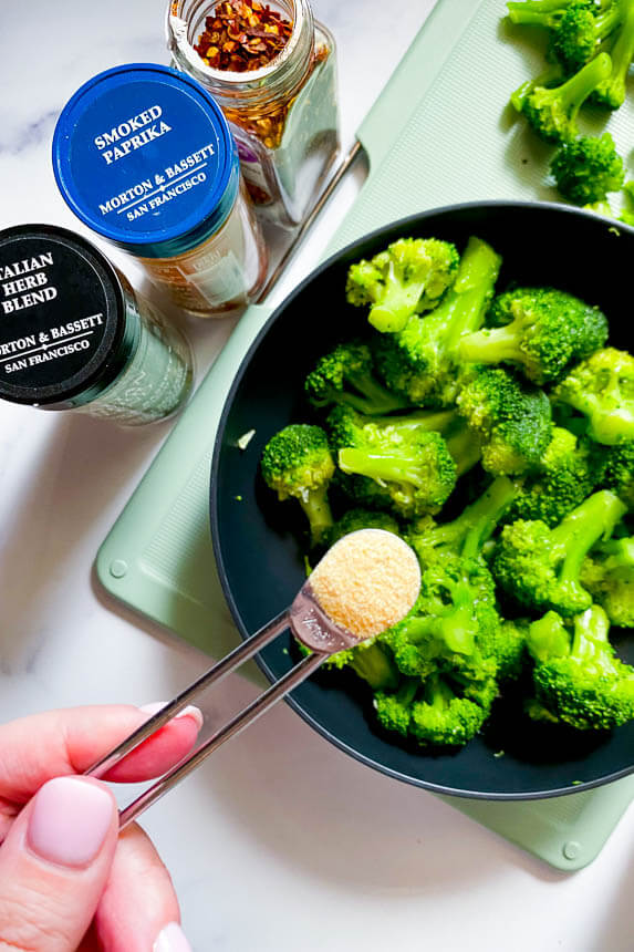 frozen broccoli in a bowl with a hand about to pour a teaspoon of garlic powder 