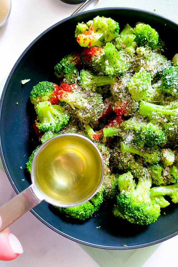 bowl of frozen broccoli sprinkled with seasonings and oil being poured 