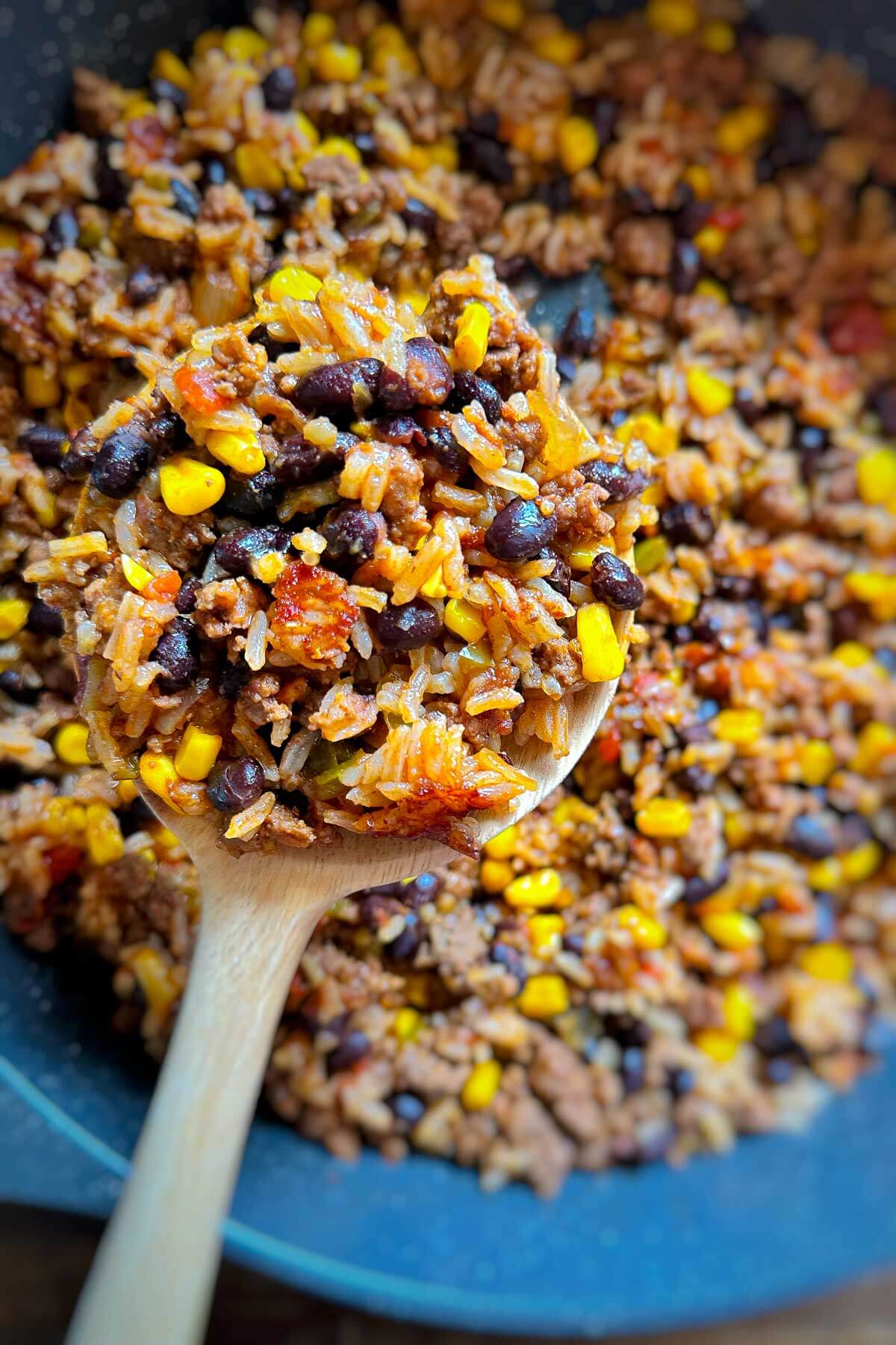 Close up of cooked taco rice with ground beef, black beans, corn, tomatoes, and seasonings scooped up with a wooden spoon from a skillet.