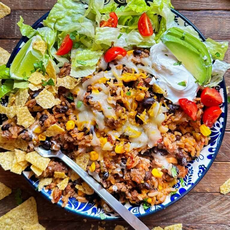 Taco rice bowl topped with melted cheese, ground beef, black beans, corn, lettuce, tomatoes, avocado, sour cream, and crushed tortilla chips on a blue patterned plate.