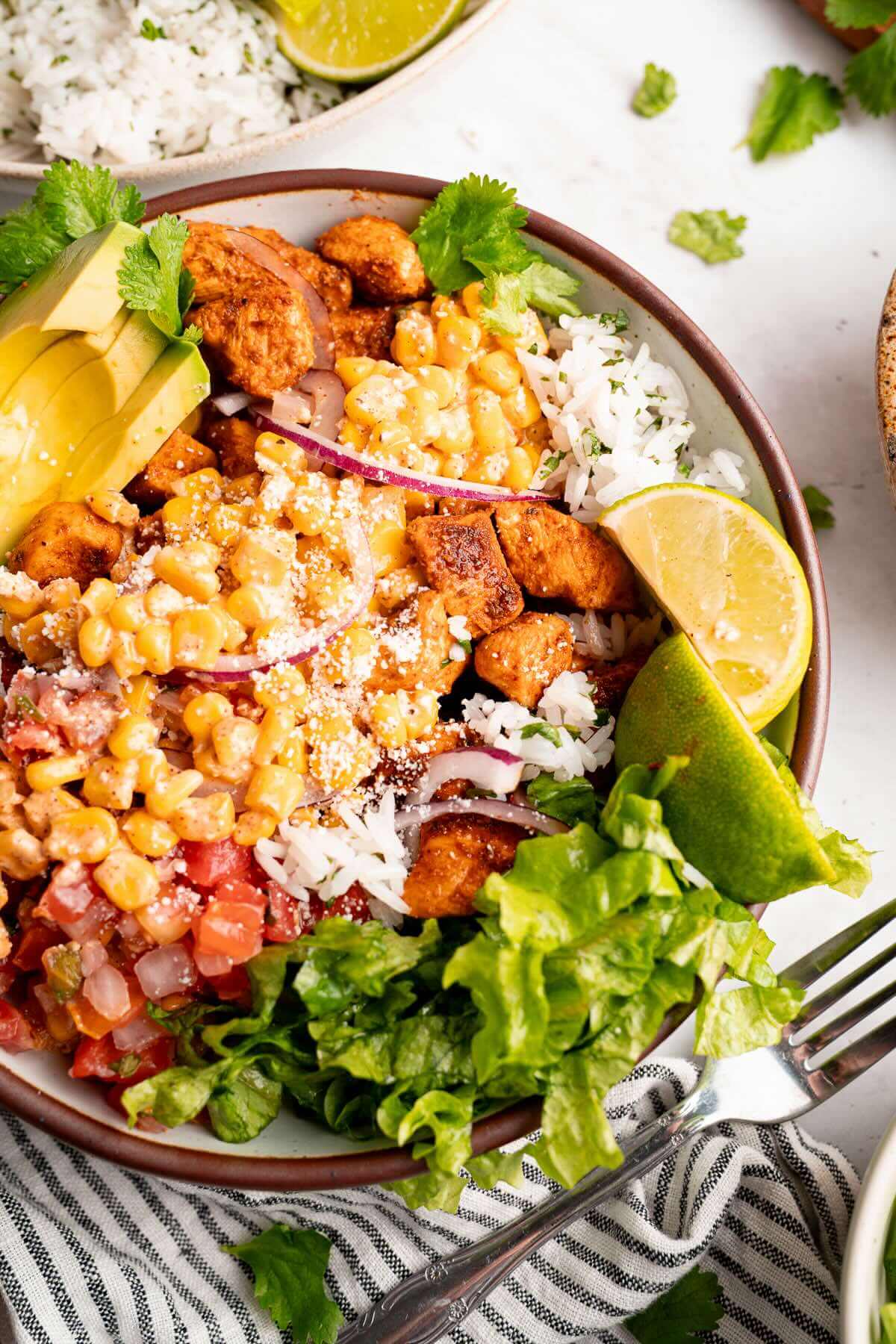 Overhead view of a street corn chicken rice bowl with seasoned chicken, corn, cilantro-lime rice, avocado, pico de gallo, lettuce, red onion, and fresh lime wedges.