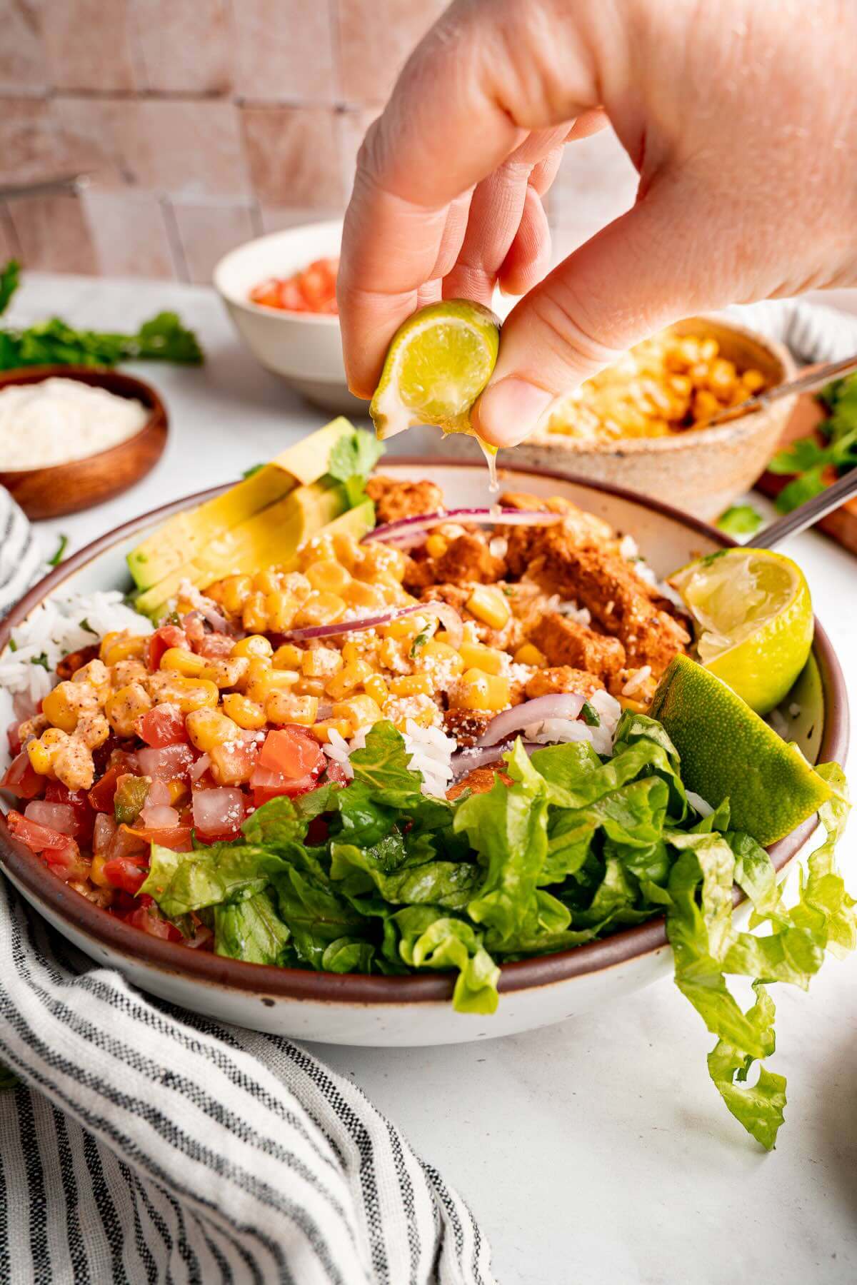 Hand squeezing fresh lime over a street corn chicken rice bowl filled with charred corn, pico de gallo, avocado, lettuce, and seasoned chicken.