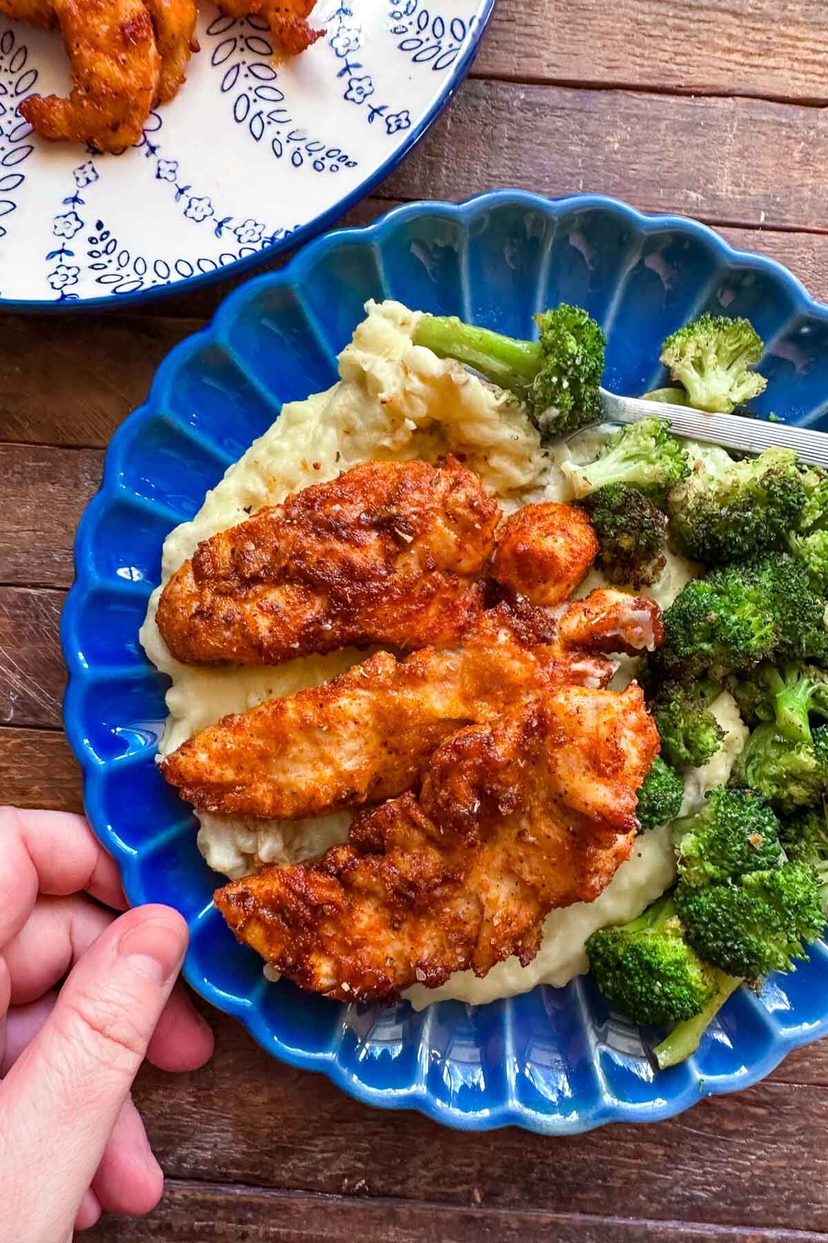 hand holding blue plate of creamy mashed potatoes, air fryer chicken tenders, and roasted broccoli 