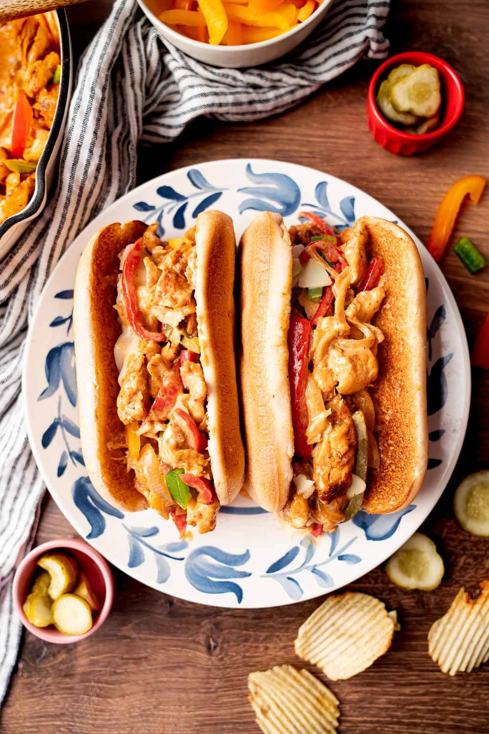 Two chicken cheesesteak sandwiches with sautéed peppers, onions, and melted cheese served with potato chips and pickles.