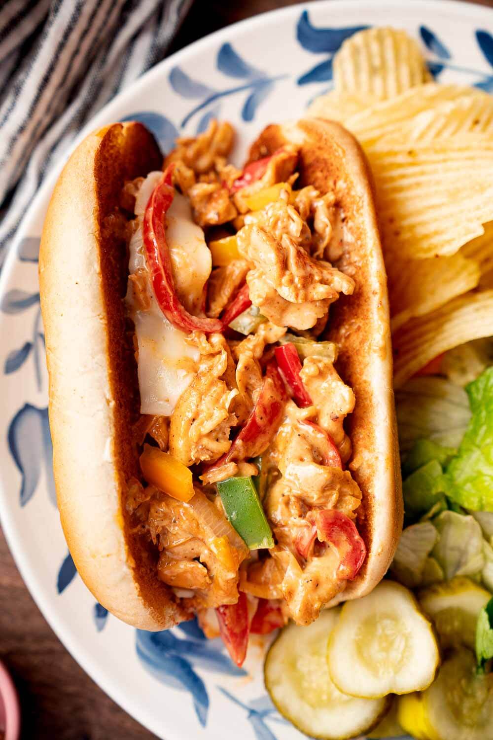 philly chicken cheesesteak sandwich on plate with chips, pickles, and salad on the side
