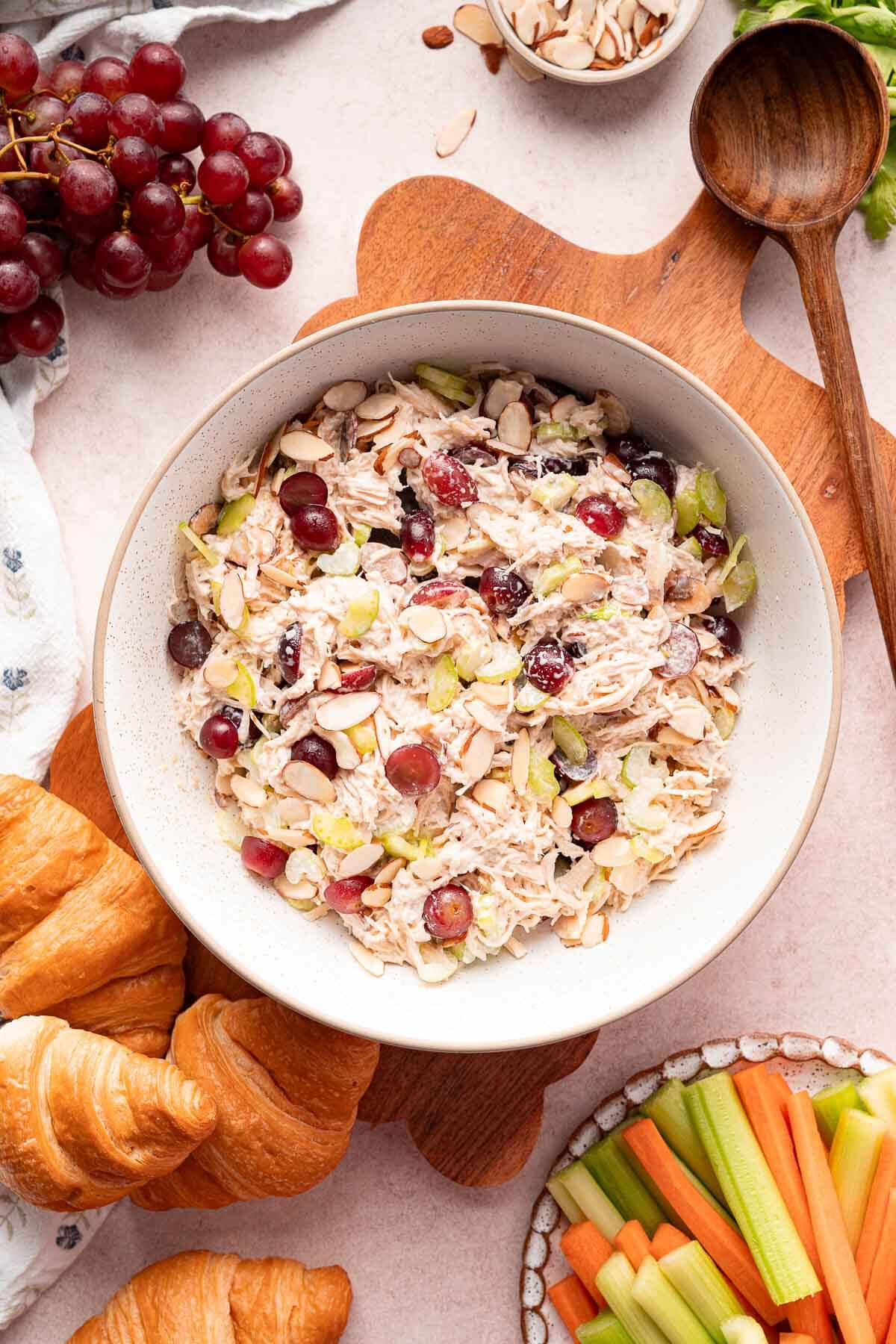 chicken salad with grapes in a big bowl surrounded y grapes, crossiants, and sliced carrots and celery. 