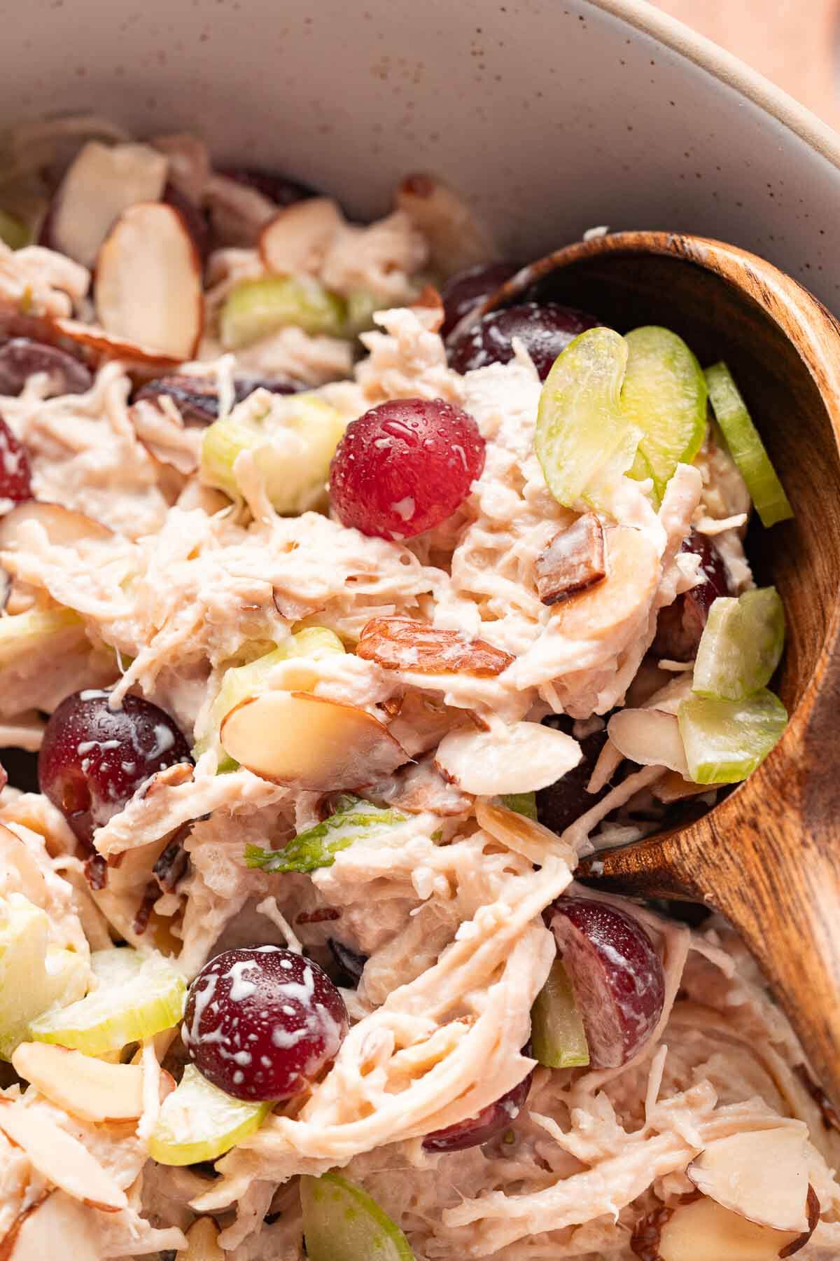 Chicken salad with grapes in a bowl with sliced almonds and celery shown in a wooden spoon.
