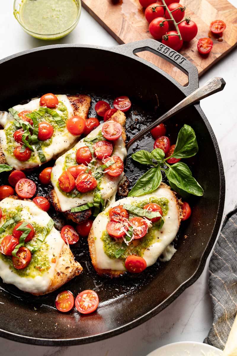 Grilled chicken margherita cooking in a skillet with melted mozzarella, basil pesto, and cherry tomatoes