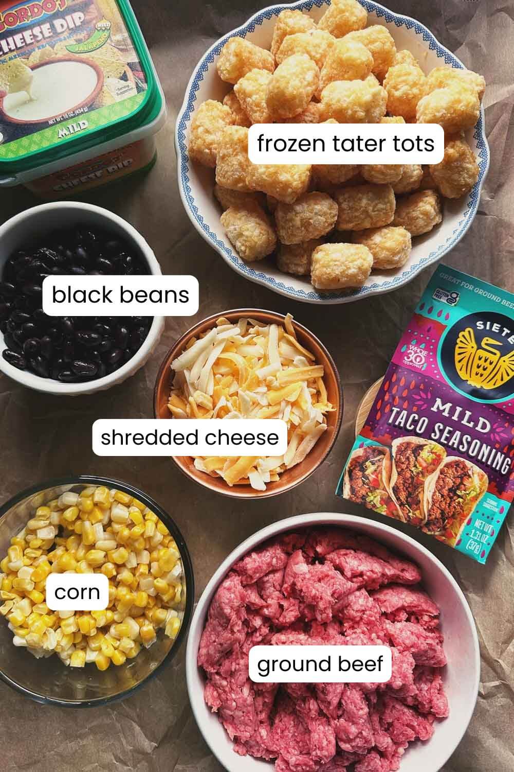 small bowls of ingredients: raw ground beef, corm, shredded cheese, black beans, frozen tater tots, a container of cheese dip, and a packet of taco seasoning.