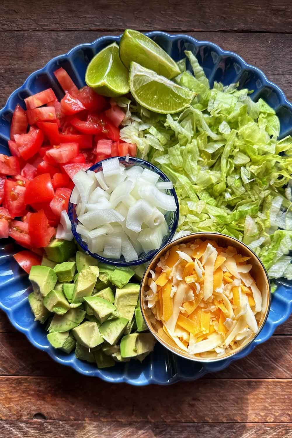 blue plate full of toppings: diced tomatoes, avocado, and onions, shredded lettuce and cheese, lime wedges