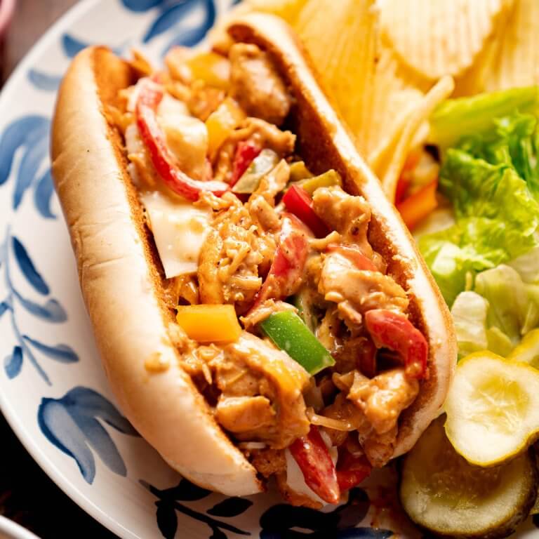 Close up of a chicken cheesesteak sandwich with sautéed peppers, onions, and melted cheese served with chips and pickles