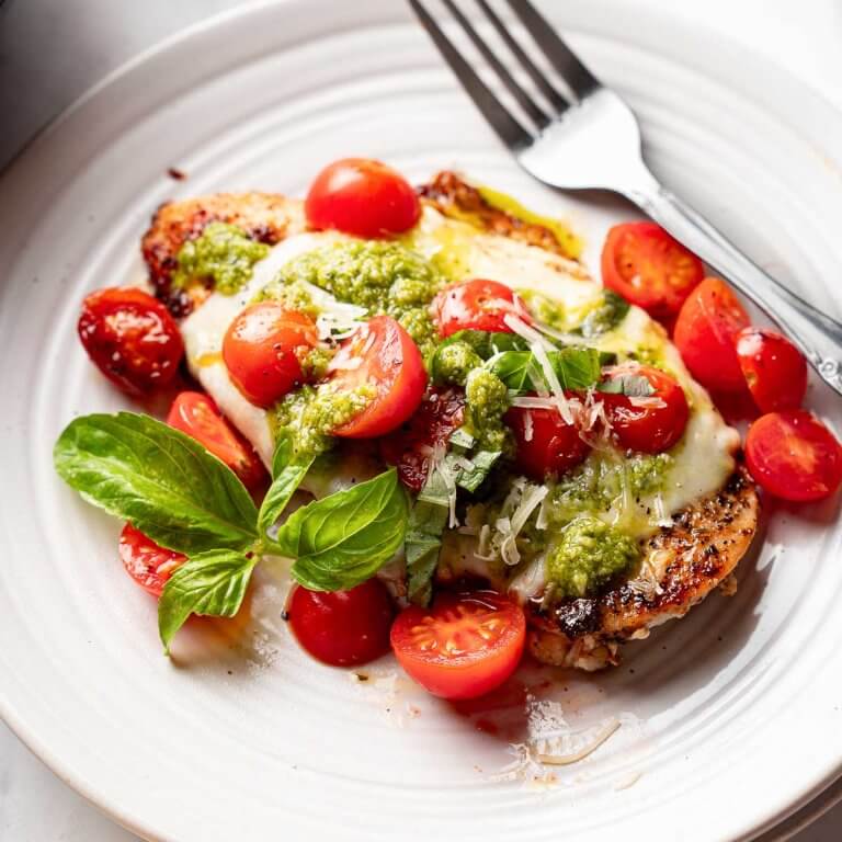 Grilled chicken margherita topped with melted mozzarella, basil pesto, and cherry tomatoes on a white plate