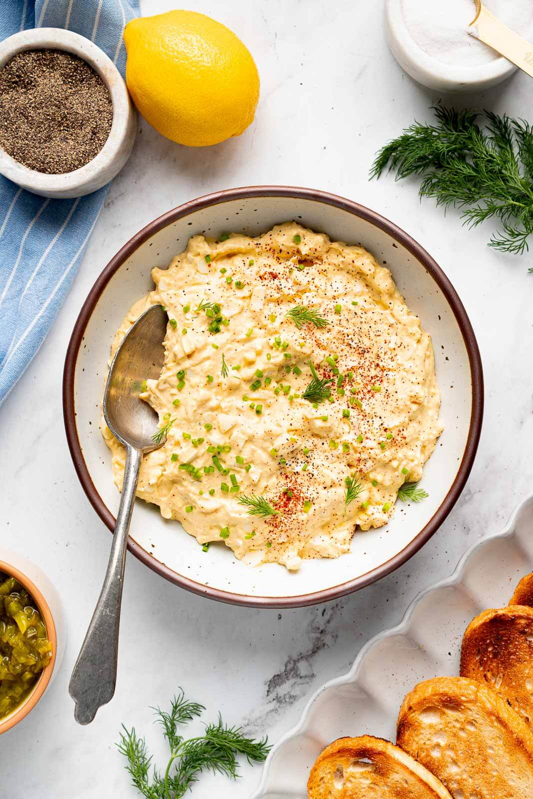 cottage cheese egg salad in bowl with spoon surrounded by toasted bread, fresh dill, and a lemon