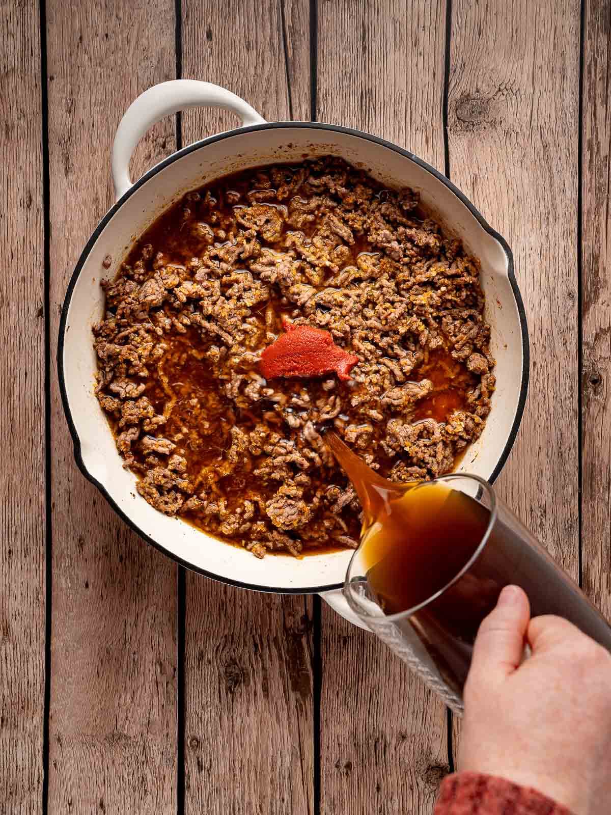 beef broth being poured into a skillet with ground beef and tomato paste.