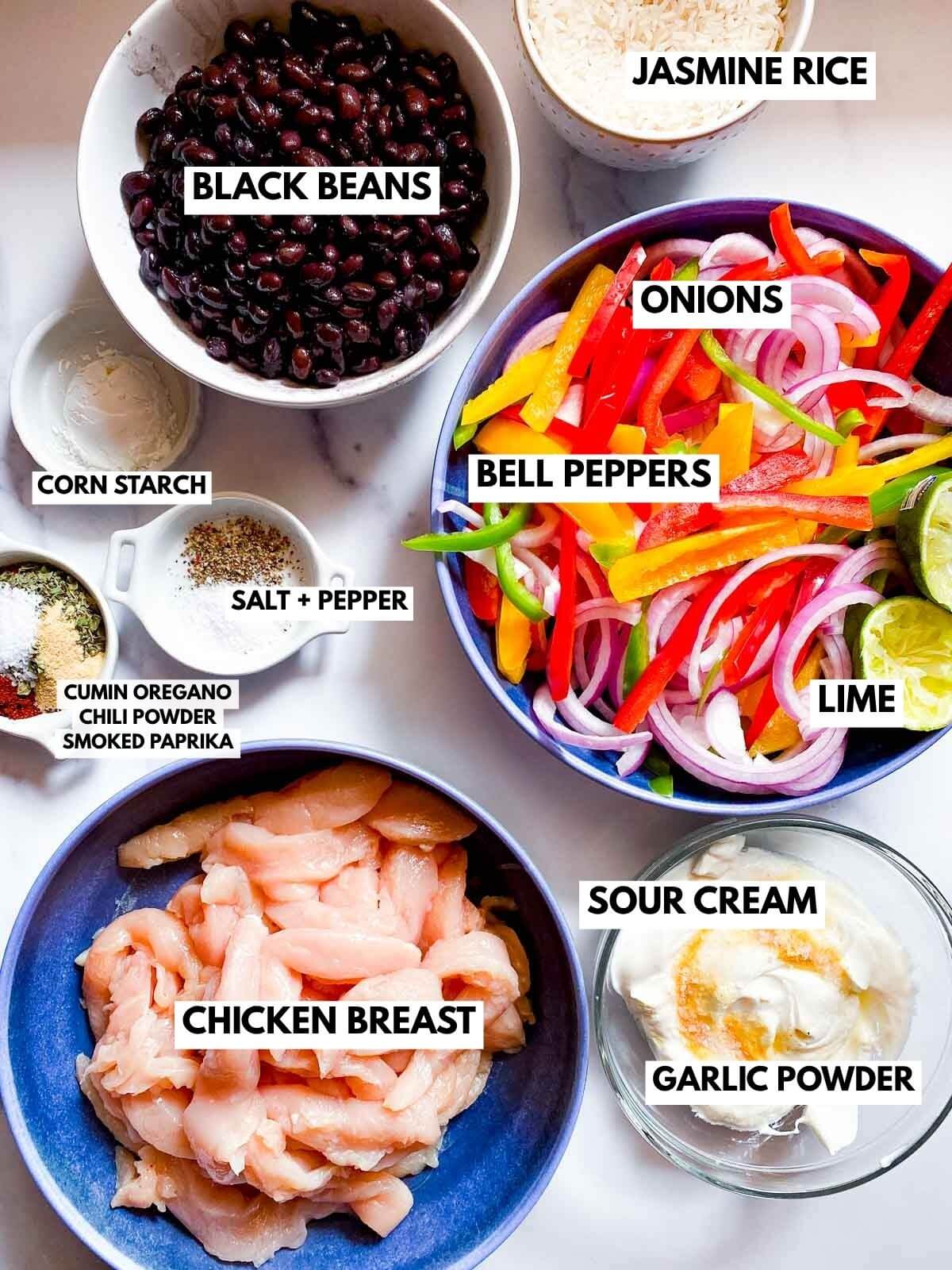 Ingredients for fajita bowls: black beans, jasmine rice, bell peppers, onions, lime, sour cream, chicken breast, corn starch, and various spices.