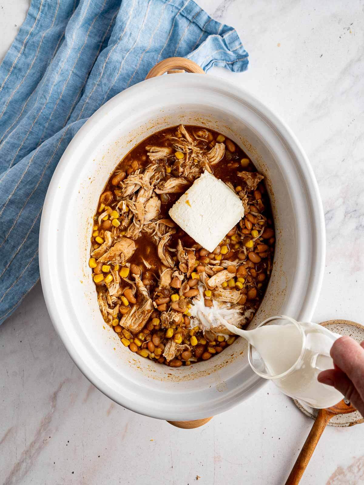 cooked and shredded chicken in crockpot with cream cheese and half and half being poured. 