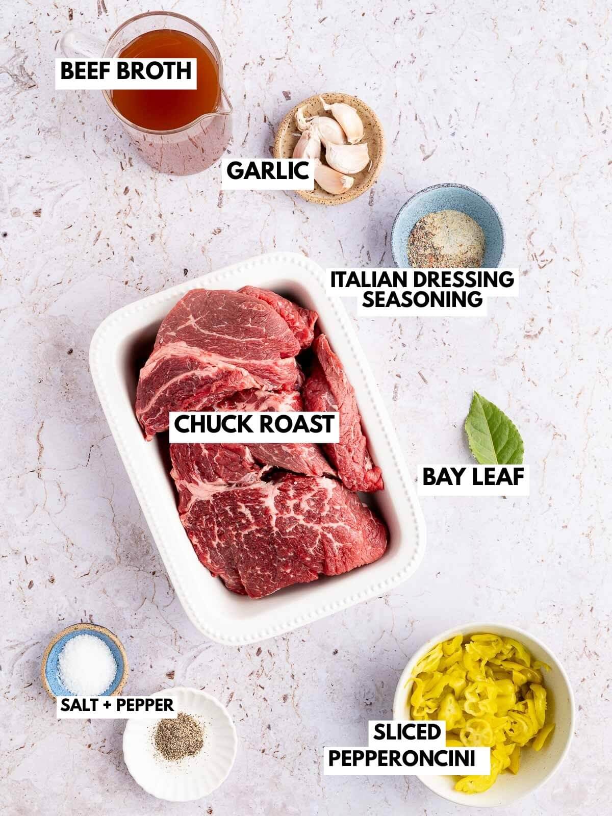 measuring cup with beef broth, small bowls with garlic  cloves, spices, pepperoncinis, salt, and pepper, chuck roast, and a bay leaf. 